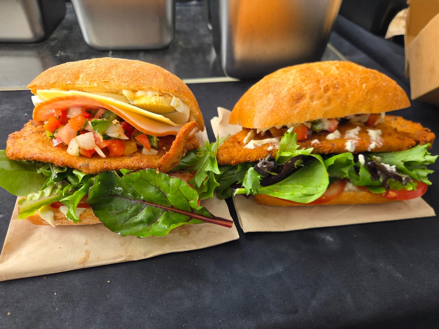 Two La Mila milanesa sandwiches — Completo and Clásico — on brown paper with fresh greens and salsa criolla